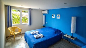 A bedroom with a blue bed, a wicker chair, a window with curtains, and a wall-mounted air conditioner. - La Cigalière Gite Bleu Azur  (ELNE)