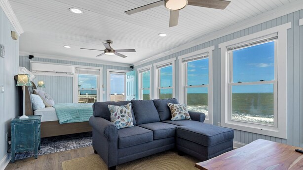 A cozy bedroom with a large bed, a gray sectional sofa, and a view of the ocean.