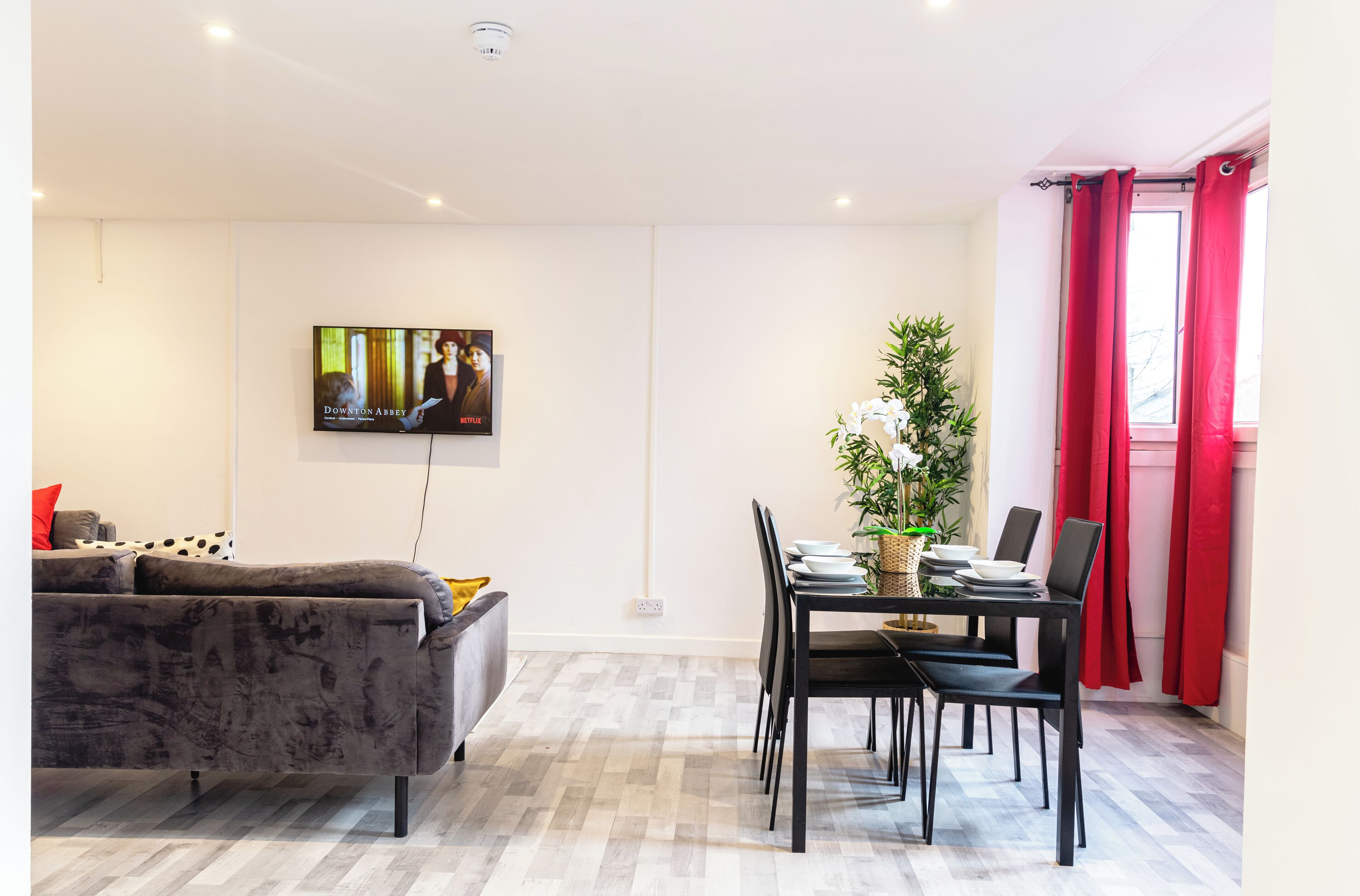 A modern living room with a grey sofa, a dining table with chairs, a potted plant, and a TV mounted on the wall.