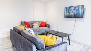 A modern living room with a grey sofa, a black coffee table, and a flat-screen TV mounted on the wall displaying a Netflix show. - 2 Bedrooms-4 Guests-babycot-wifi-freeparking (London)