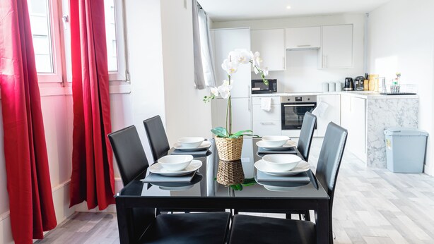 A modern dining area with a glass table set for four, surrounded by black chairs. A woven basket with white orchids is placed on the table. - 2 Bedrooms-4 Guests-babycot-wifi-freeparking (London)