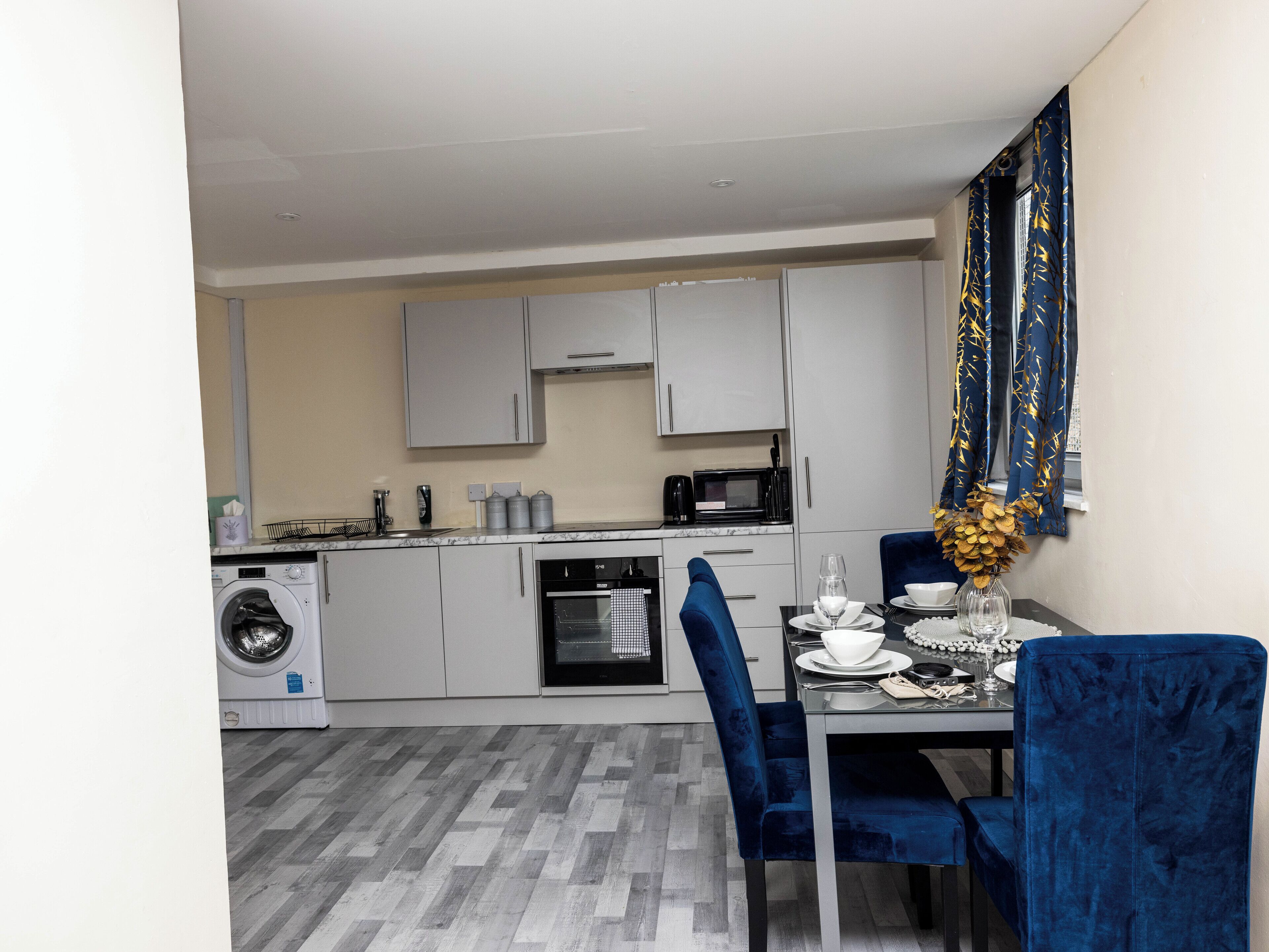 A compact kitchen with a washing machine, microwave, and oven. There is a dining area with a table set for two and blue chairs.