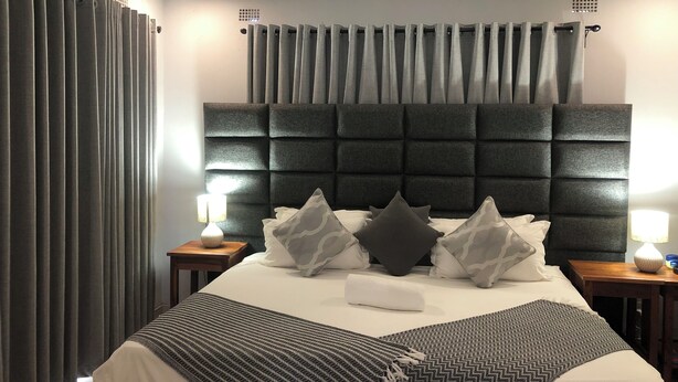 Una habitación de hotel moderna con una cama grande, cortinas grises, un cabecero oscuro y dos lámparas de noche.