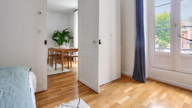 A room with wooden flooring, a bed, a dining area with a table and chairs, and a large window with a view of buildings and trees. - Flat in the town centre - 270m  l'Ecluse beach (Dinard)