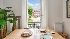A dining table set with plates, glasses, and a bottle, with a view of a red building and a tree outside. - Flat in the town centre - 270m  l'Ecluse beach (Dinard)