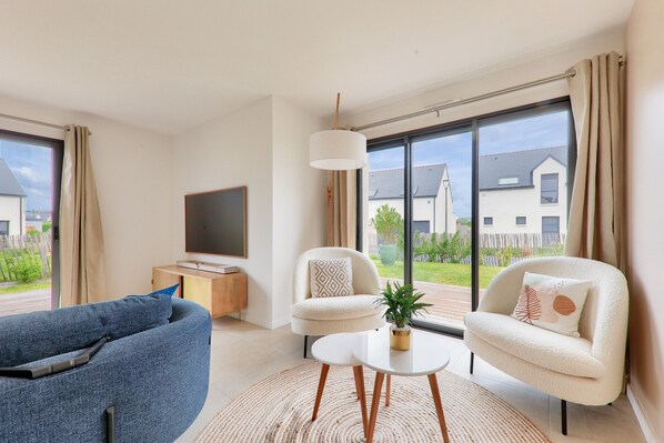 A modern living room with a sofa, two armchairs, a coffee table, a TV, and large windows with a view of houses.