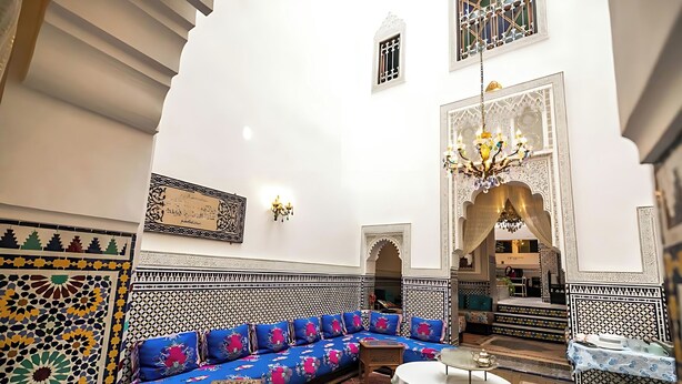 A traditional Moroccan interior with a patterned sofa, a chandelier, and a view of the interior through an archway. - Riad Mounana luxurious palace & spa  (Fes)