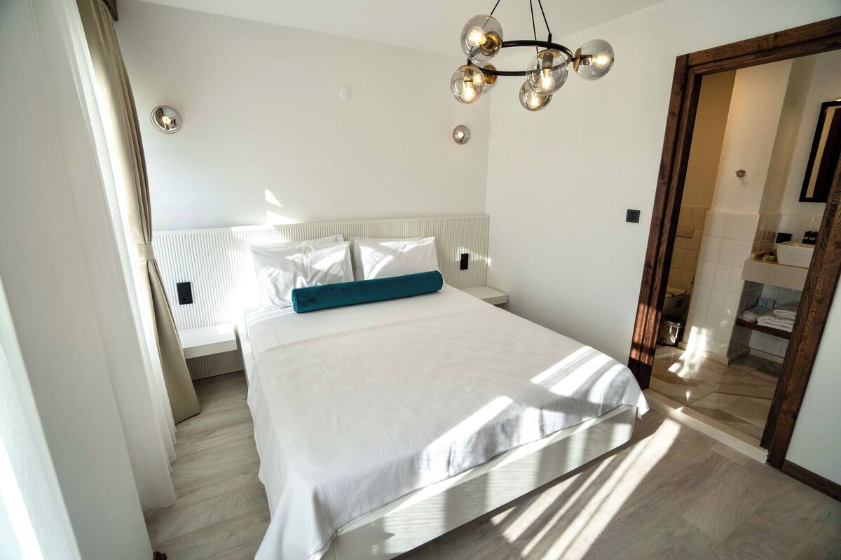 A neatly made bed with a white and teal pillow, a modern chandelier, and a view into the bathroom through an open door.