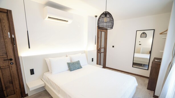 A bedroom with a large bed, a wooden wardrobe, a hanging light fixture, and a mirror on the wall. - Lina Guest House (Ayvalik)