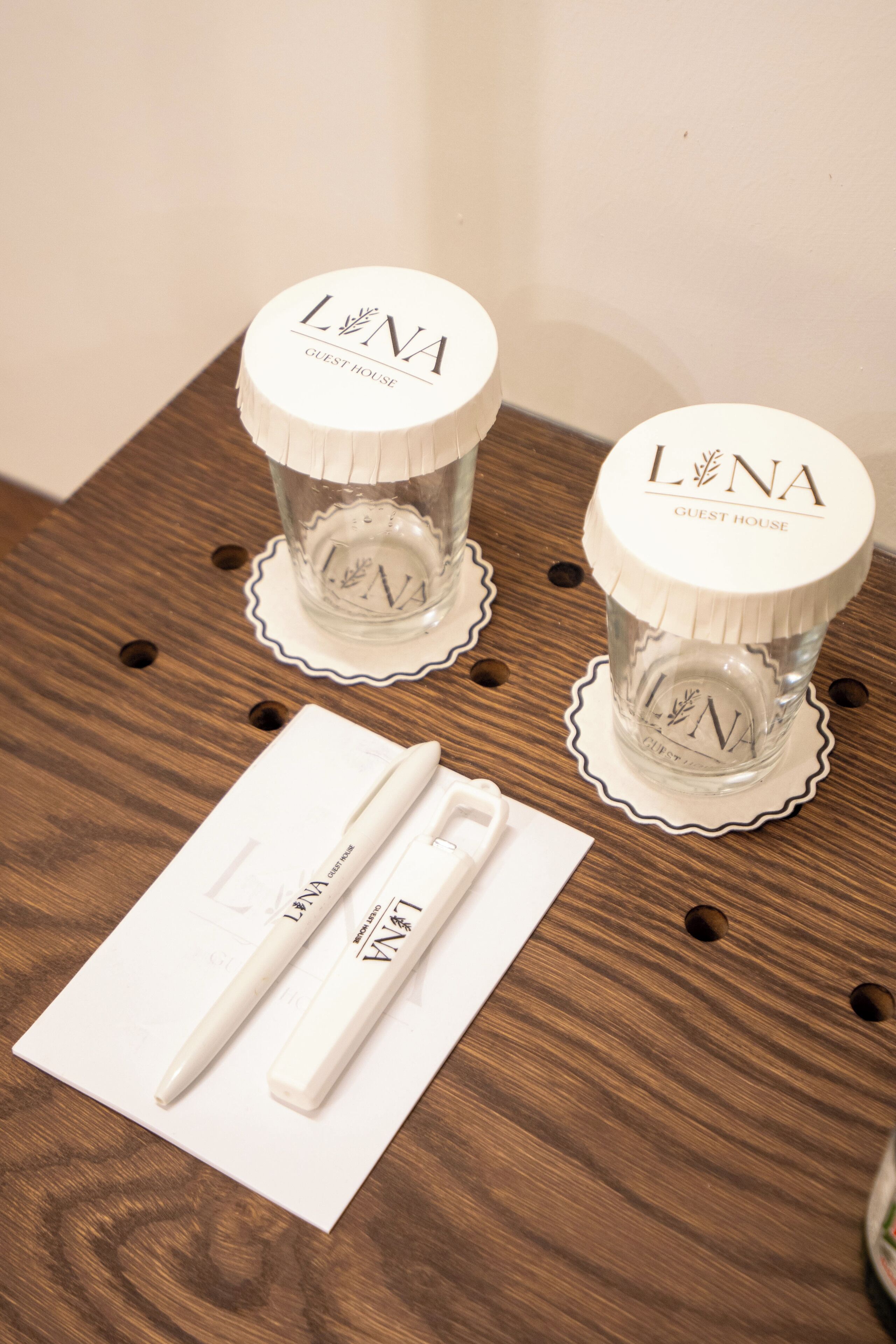 Two glass containers with lids on a wooden table, each labeled "L'NA GUEST HOUSE".