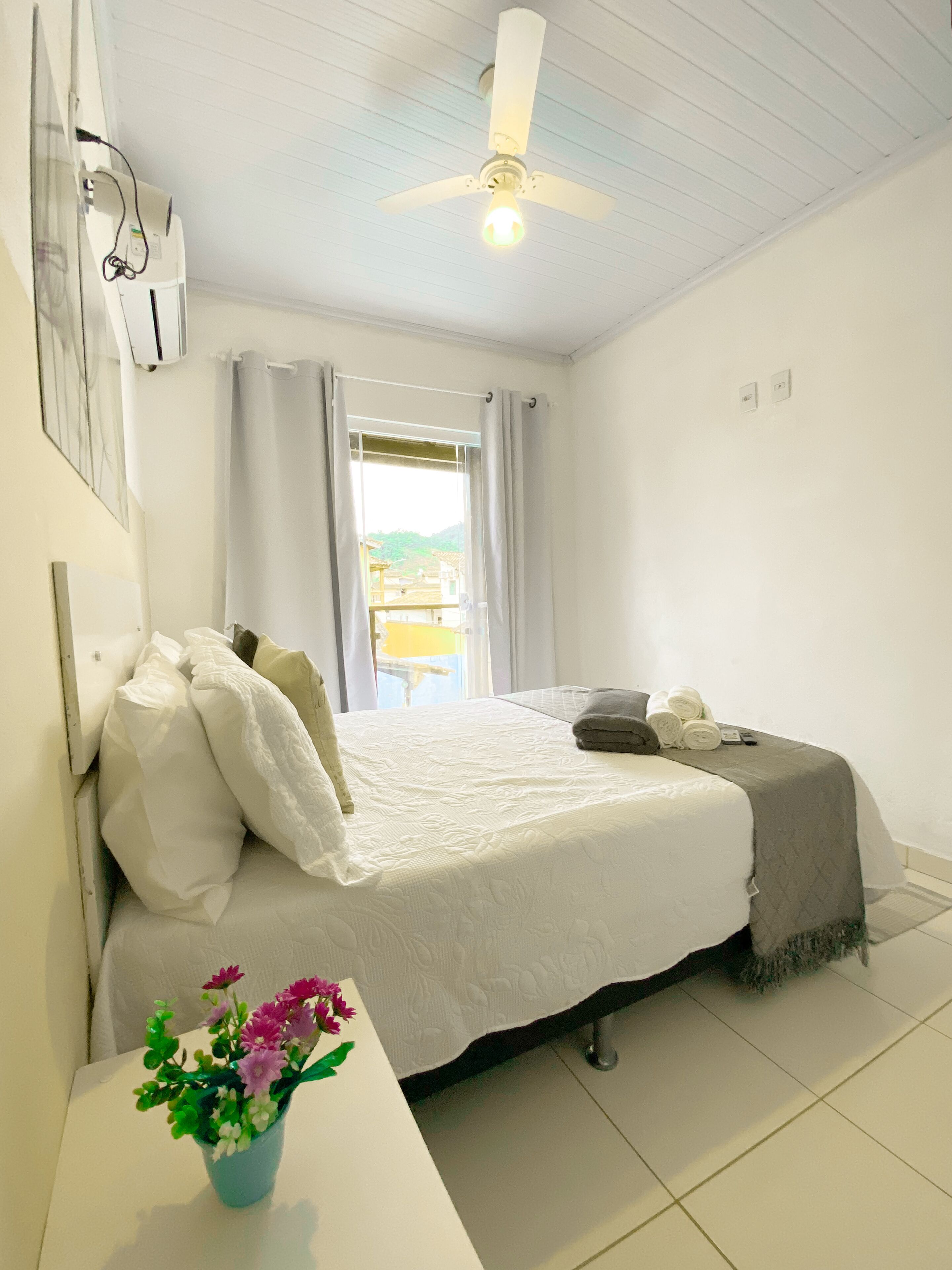 Un dormitorio con cama, una mesita de noche con un florero, un ventilador de techo y una ventana con cortinas.
