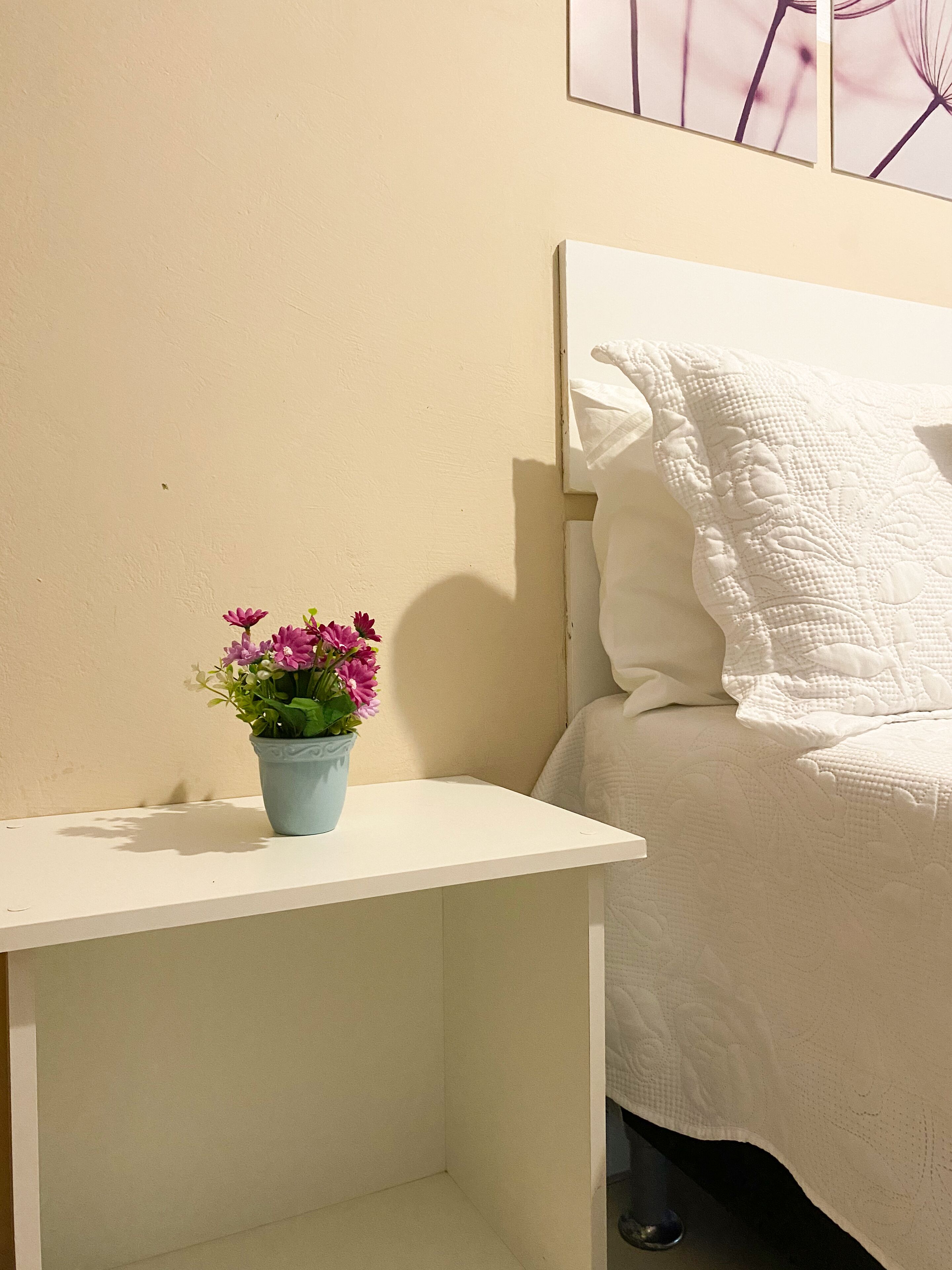 A white bedside table with a blue pot containing pink flowers, a white bed with a textured blanket, and a wall with a framed picture.