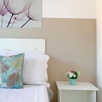 Una cama bien hecha con ropa blanca y una almohada floral, una mesita de noche pequeña de color blanco con una planta en maceta, y una obra de arte en la pared sobre la cama.