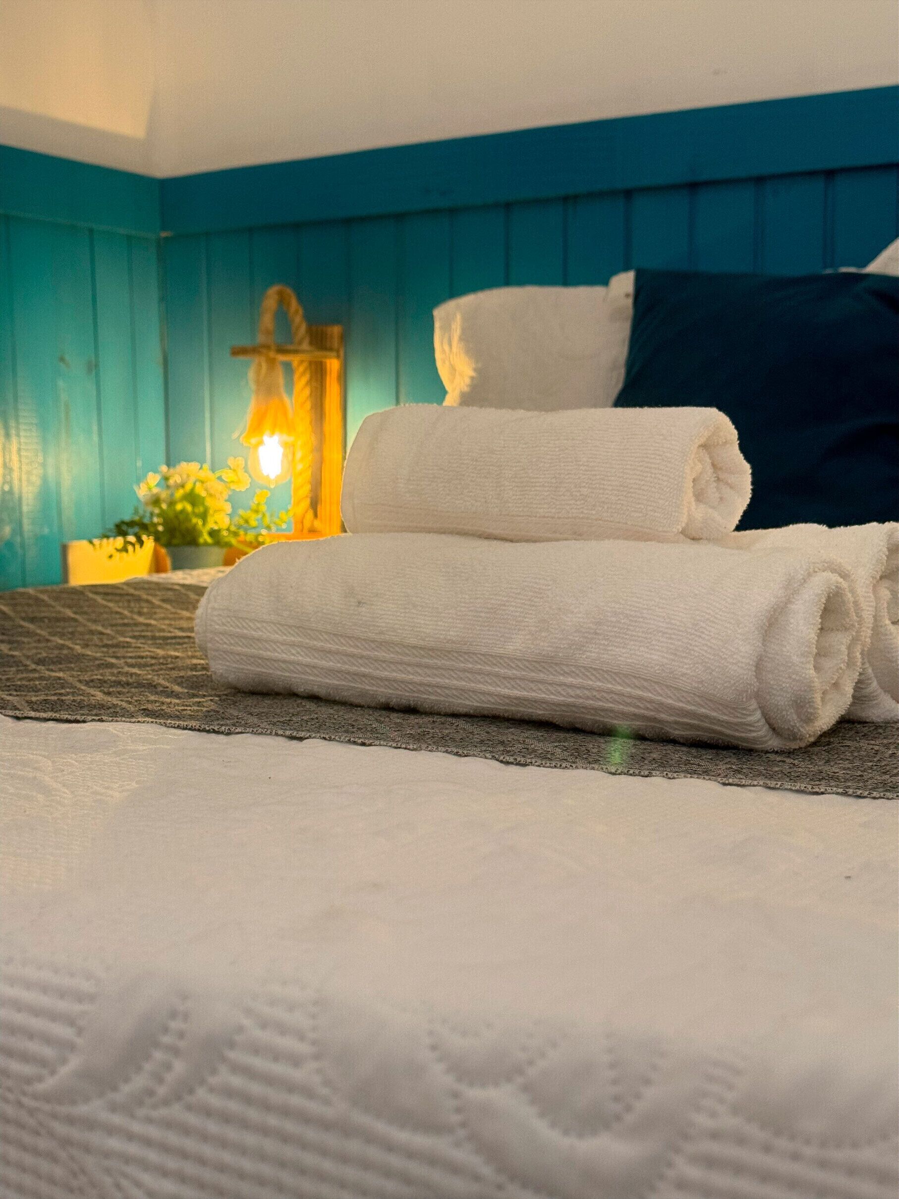 A neatly made bed with rolled white towels, a dark blue headboard, and a bedside lamp with a yellow shade.