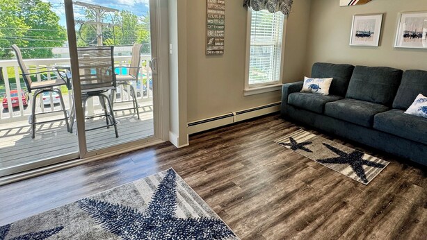 A living room with a grey sofa, a patterned rug, a wooden floor, and a view of a pool and cars through a sliding glass door. - Updated Condo at The Lookout in Ogunquit (Ogunquit)