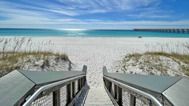 Une plage avec une jetée visible au loin, un ciel dégagé et un sentier de sable menant à l’eau.