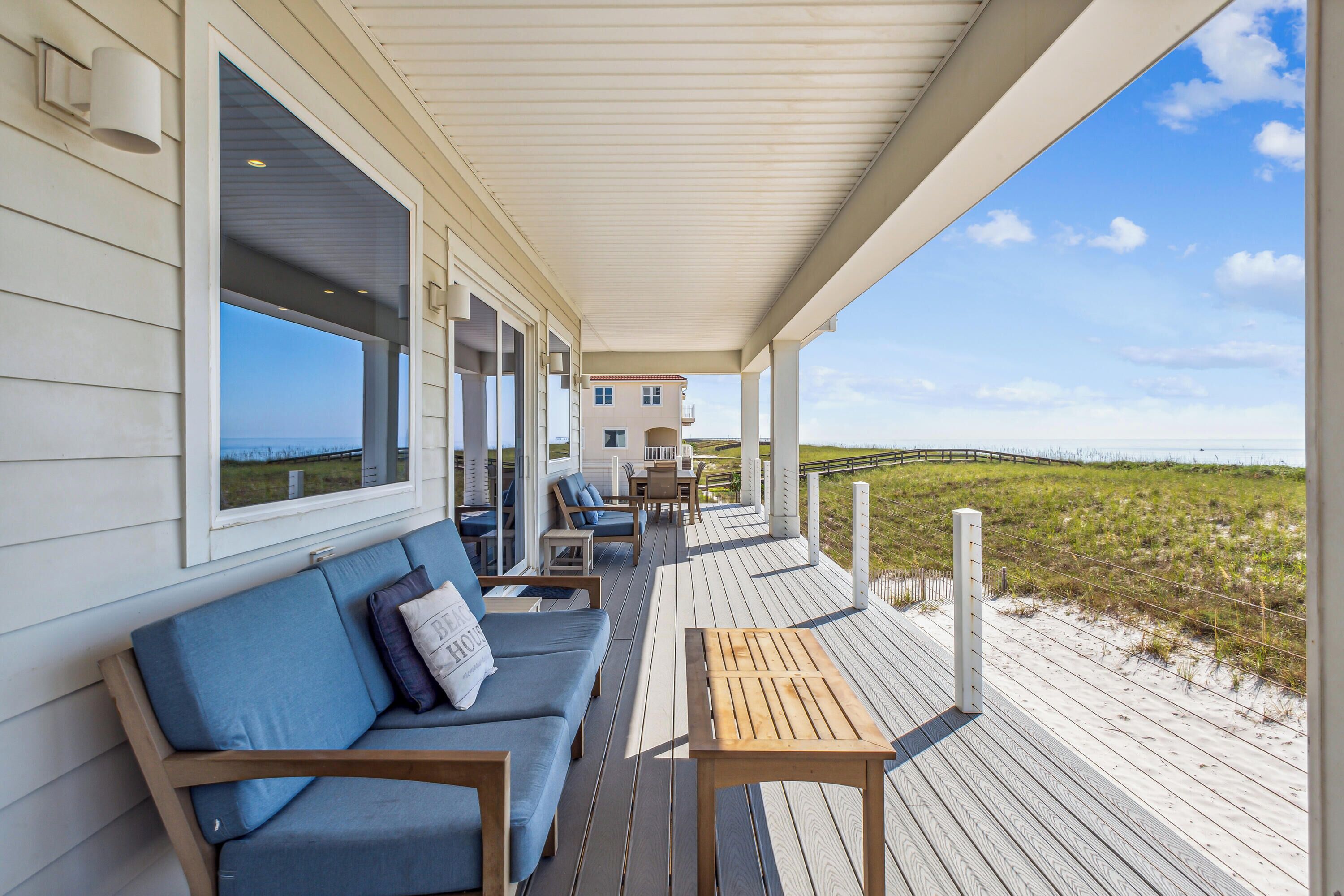 A covered deck with a sofa and table, overlooking a beach and dunes.