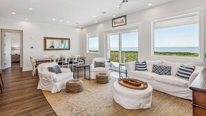 Un salon spacieux doté d’une grande fenêtre donnant sur la plage, d’un ventilateur de plafond et d’un coin repas avec des meubles blancs.