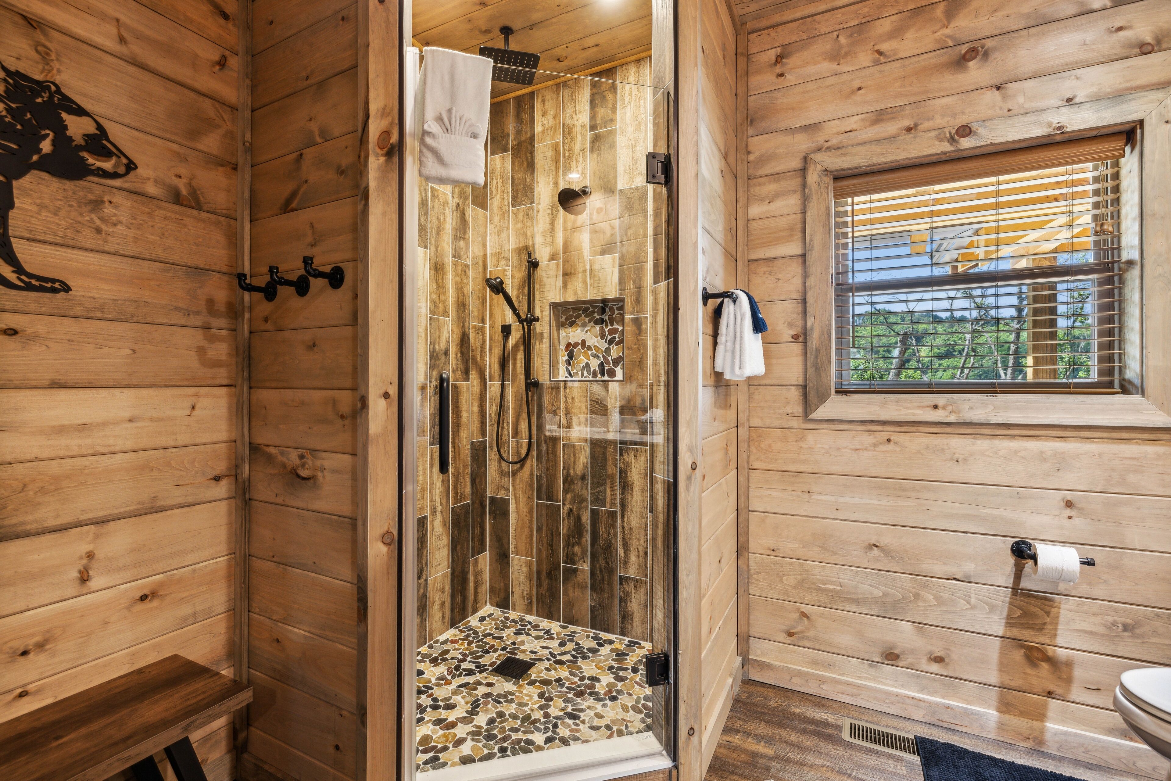 A wooden cabin bathroom with a glass-enclosed shower, wooden walls, a window with blinds, and a toilet.