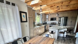 A spacious kitchen with wooden cabinets and a large island, a dining area with a wooden table and chairs, and a view of the outdoors through a window. - Eva's Place Limit 7 4 Bedroom Home by RedAwning (Sebago)