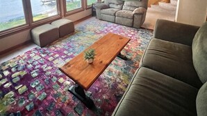 Interior de una cabaña con techo de madera, ventanas amplias con vista a un lago, un sofá, una mesa de centro y una escalera.