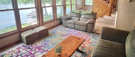 A cabin interior with a wooden ceiling, large windows overlooking a lake, a sofa, a coffee table, and a staircase.