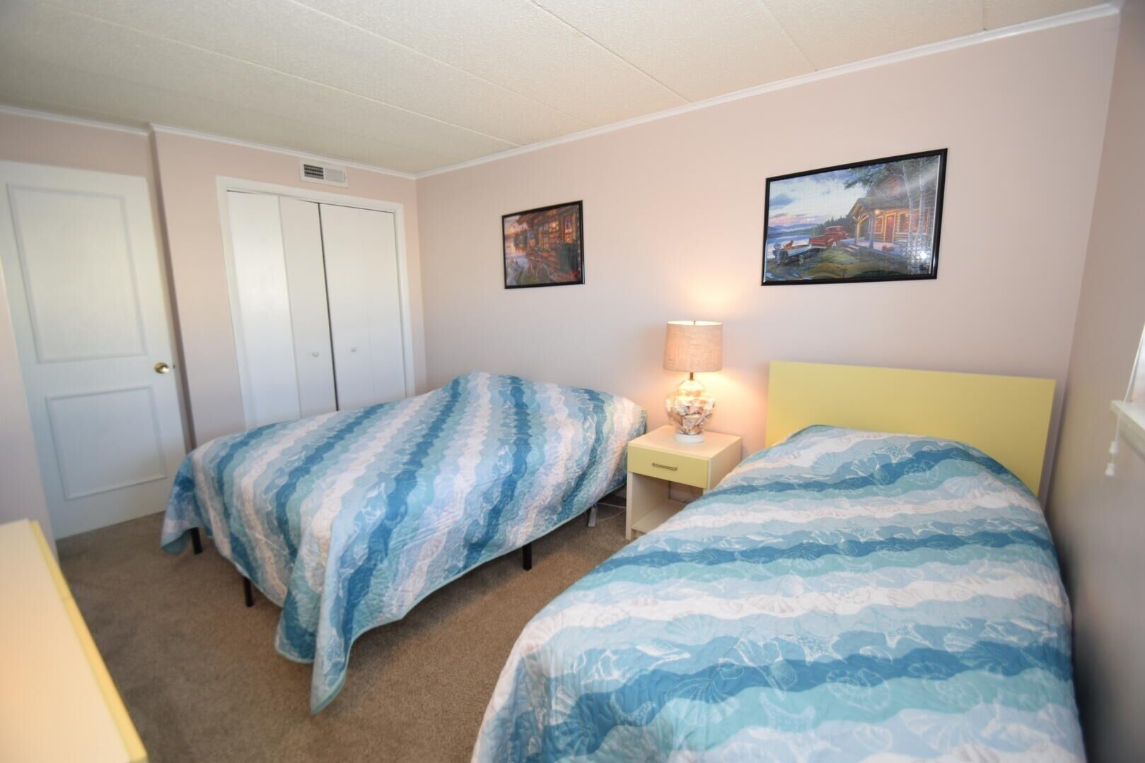 Un dormitorio con dos camas individuales, una mesita de noche con lámpara y dos cuadros en la pared.