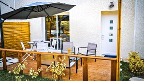 An outdoor patio with a wooden table and chairs, a black umbrella, and a view of the interior through glass doors. - La Cigalière Gite Rouge Cerise Avec Piscine et Salle de Sport Proche St Cyprien (ELNE)