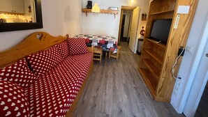Une pièce chaleureuse avec un canapé doté d’une tête de lit en bois, une petite table recouverte d’une nappe à carreaux, un meuble en bois sur lequel est posée une télévision, et un miroir accroché au mur.