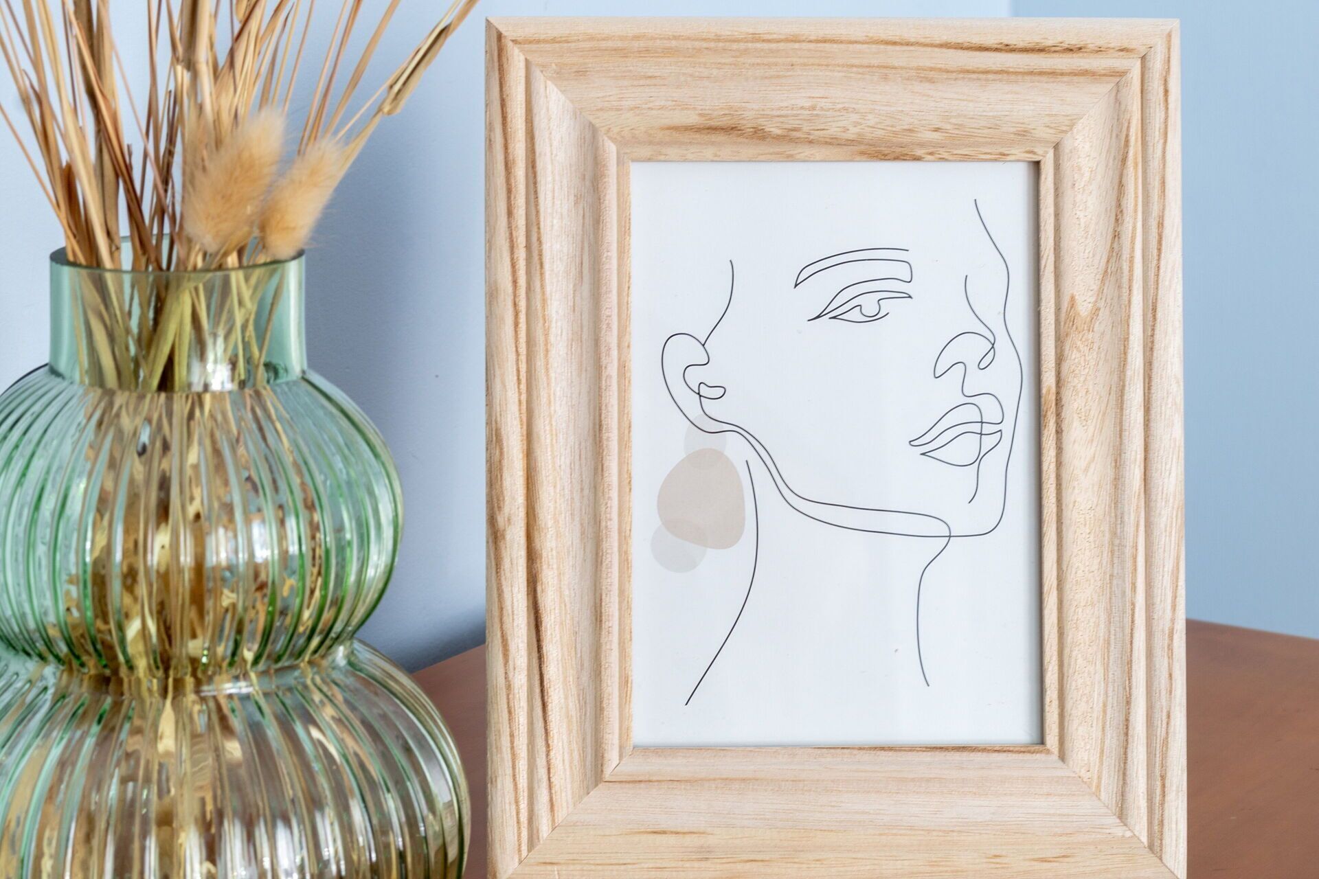 A framed line drawing of a face, a glass vase with dried plants, and a wooden table.