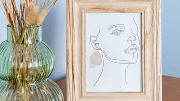 A framed line drawing of a face, a glass vase with dried plants, and a wooden table.