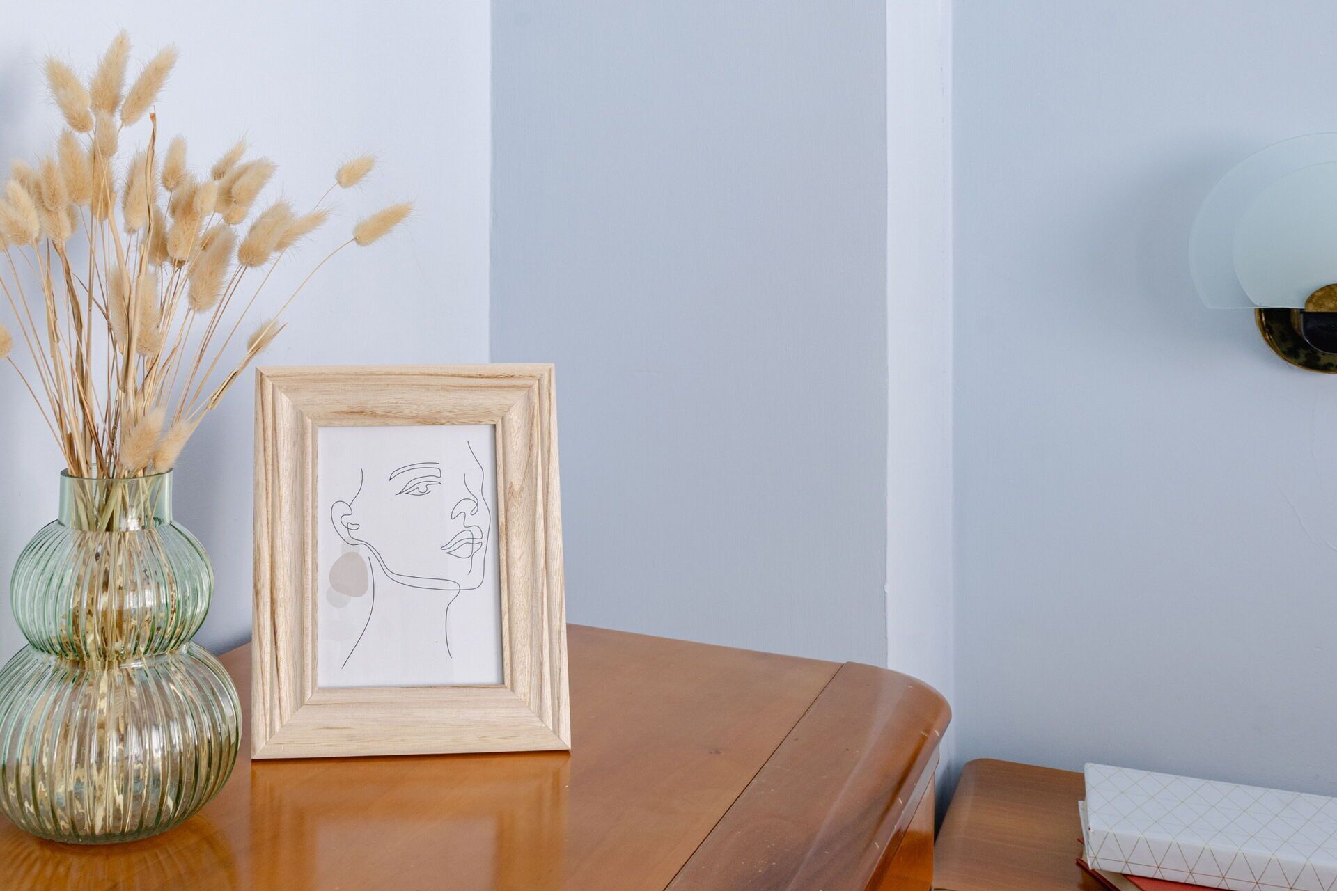 A framed line drawing of a face on a wooden table next to a glass vase with dried plants.