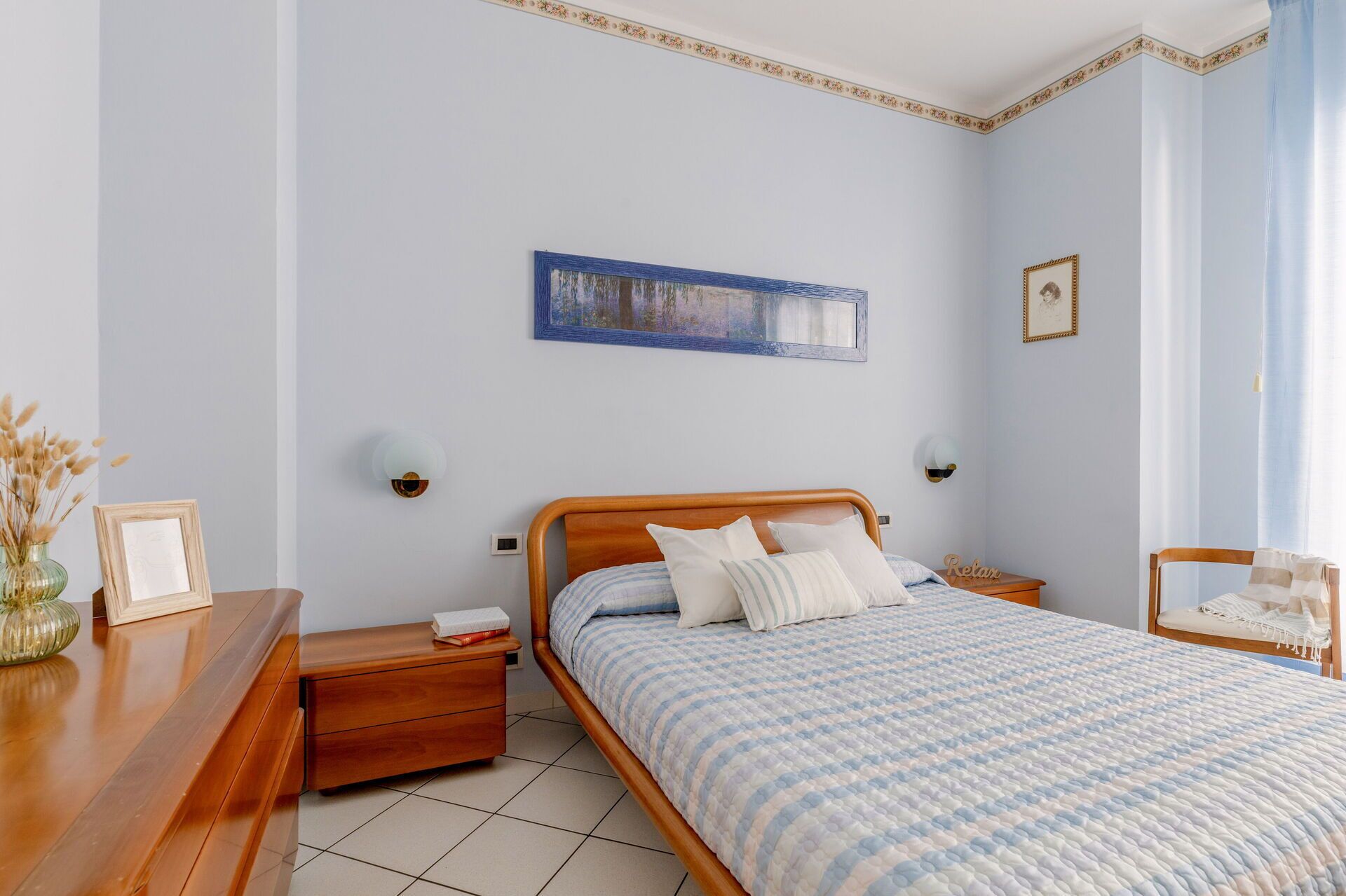 A bedroom with a wooden bed, a nightstand, a bedside table, a wall-mounted lamp, and a framed picture.