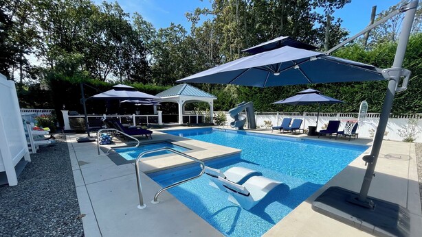 A swimming pool with lounge chairs and umbrellas, surrounded by a white fence and trees. - 6,000 Sq.ft. 18 Guests, 7-Bed, 6-Bath, Pool, Spa, Outdoor Kitchen, Garden, WIFI. (Middle Township)