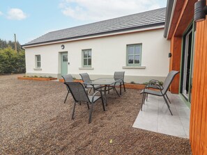 A building with a white exterior, a green door, and a window with a green frame. There is a gravel patio with a table and chairs.