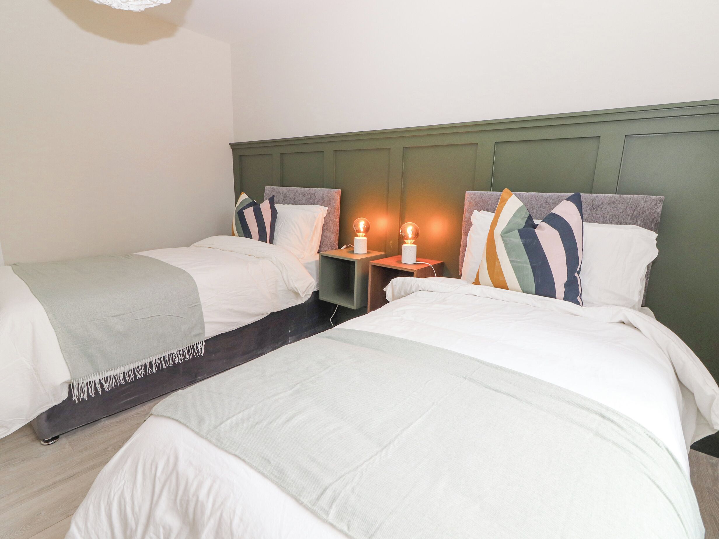 A hotel room with two beds, each with white bedding and striped pillows. There are bedside tables with lamps and a small nightstand.