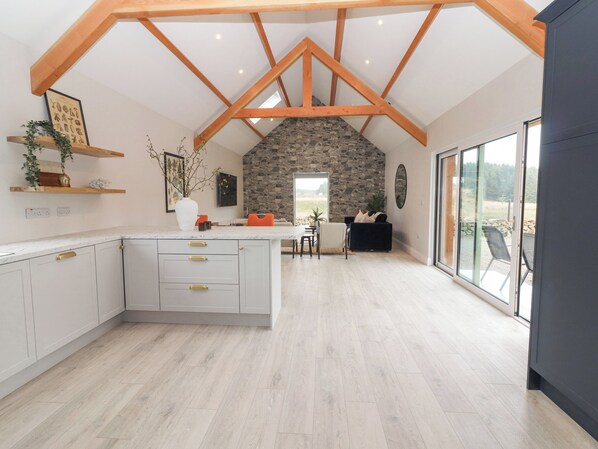 A spacious kitchen with a central island, wooden beams, stone wall, and large windows offering a view of the outdoors.