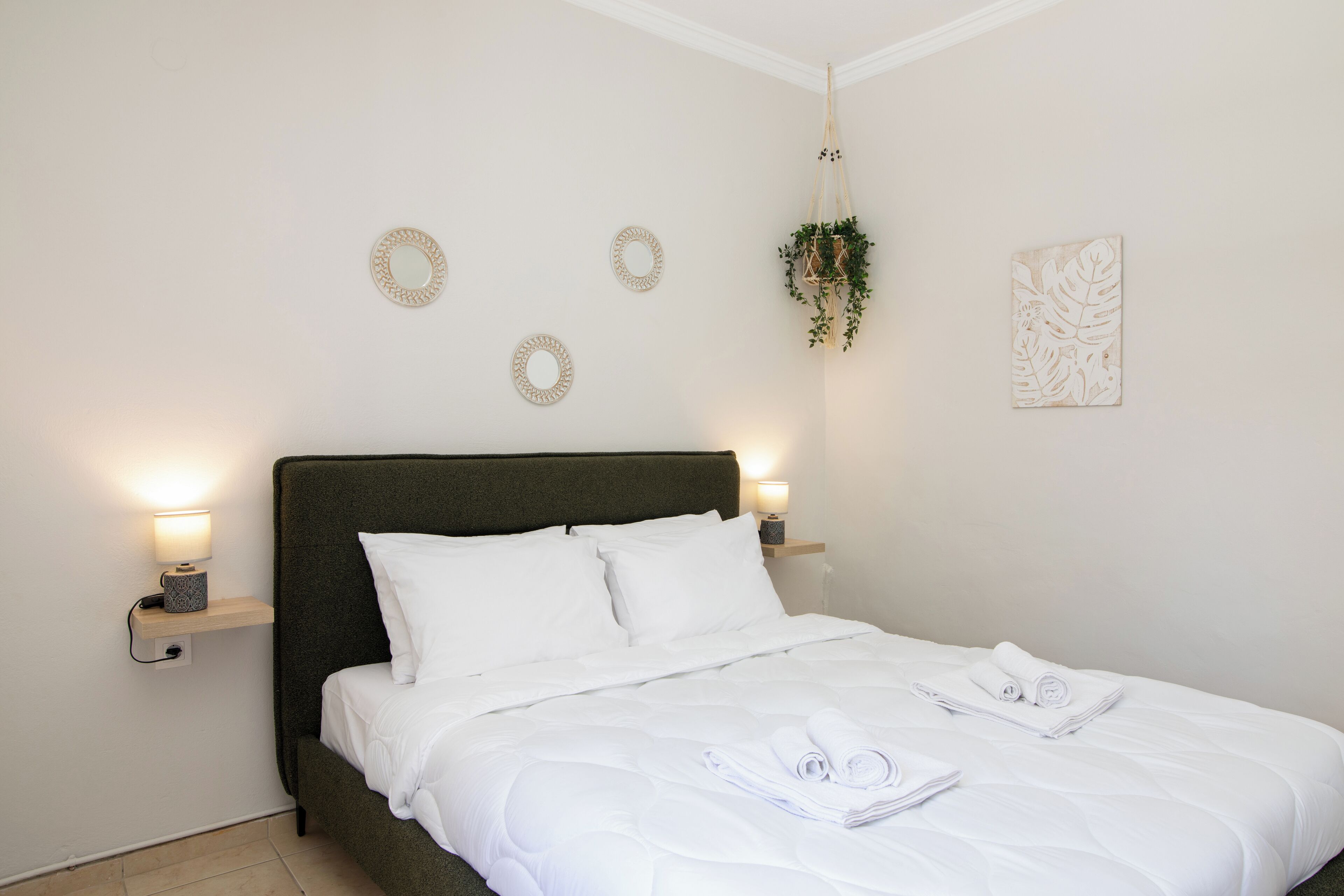 Un lit bien fait, avec du linge de lit blanc et des serviettes pliées, une tête de lit vert foncé et des éléments de décoration fixés au mur, notamment des miroirs ronds et une plante.