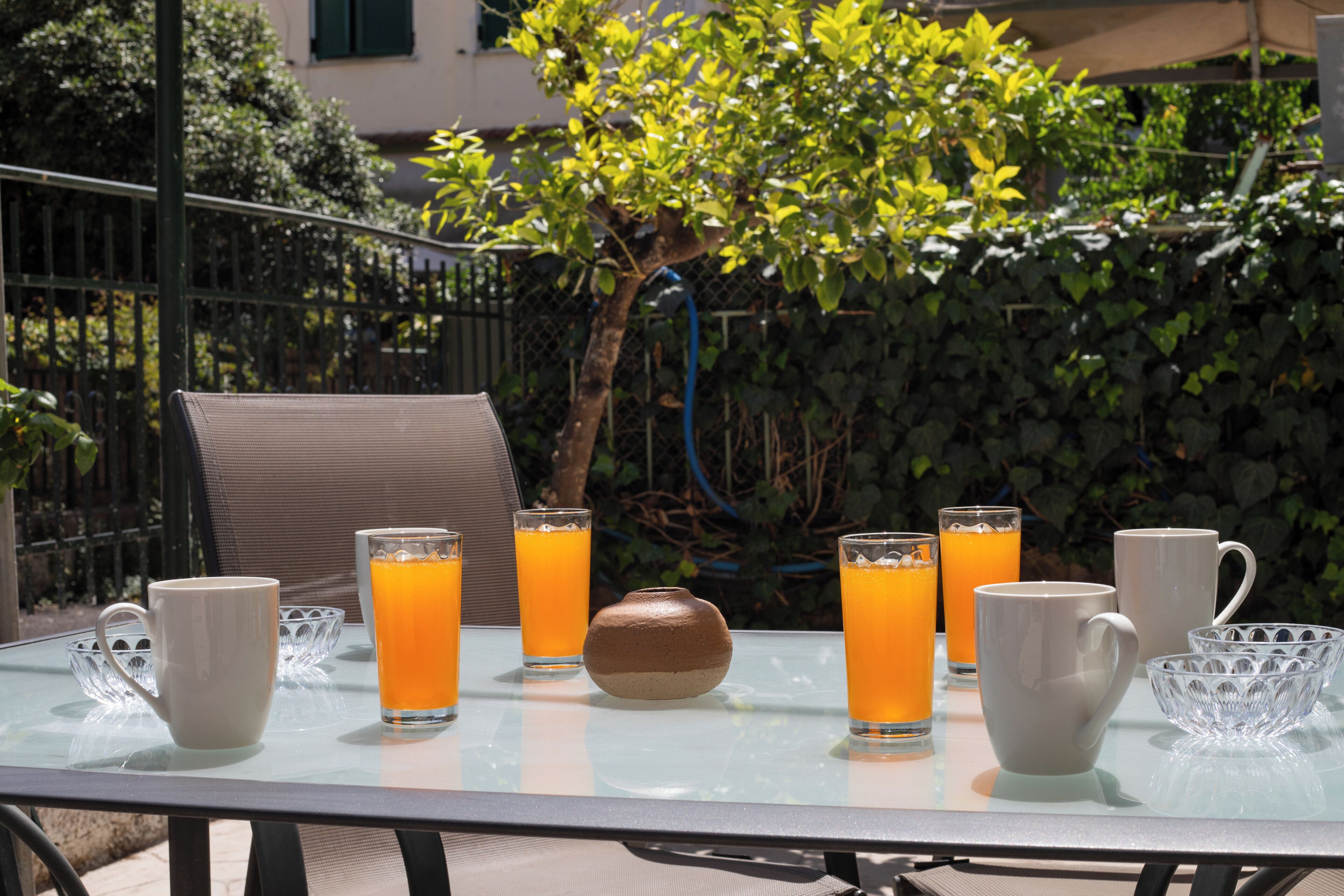 Una mesa con cuatro vasos de zumo de naranja, una taza blanca y un florero decorativo.