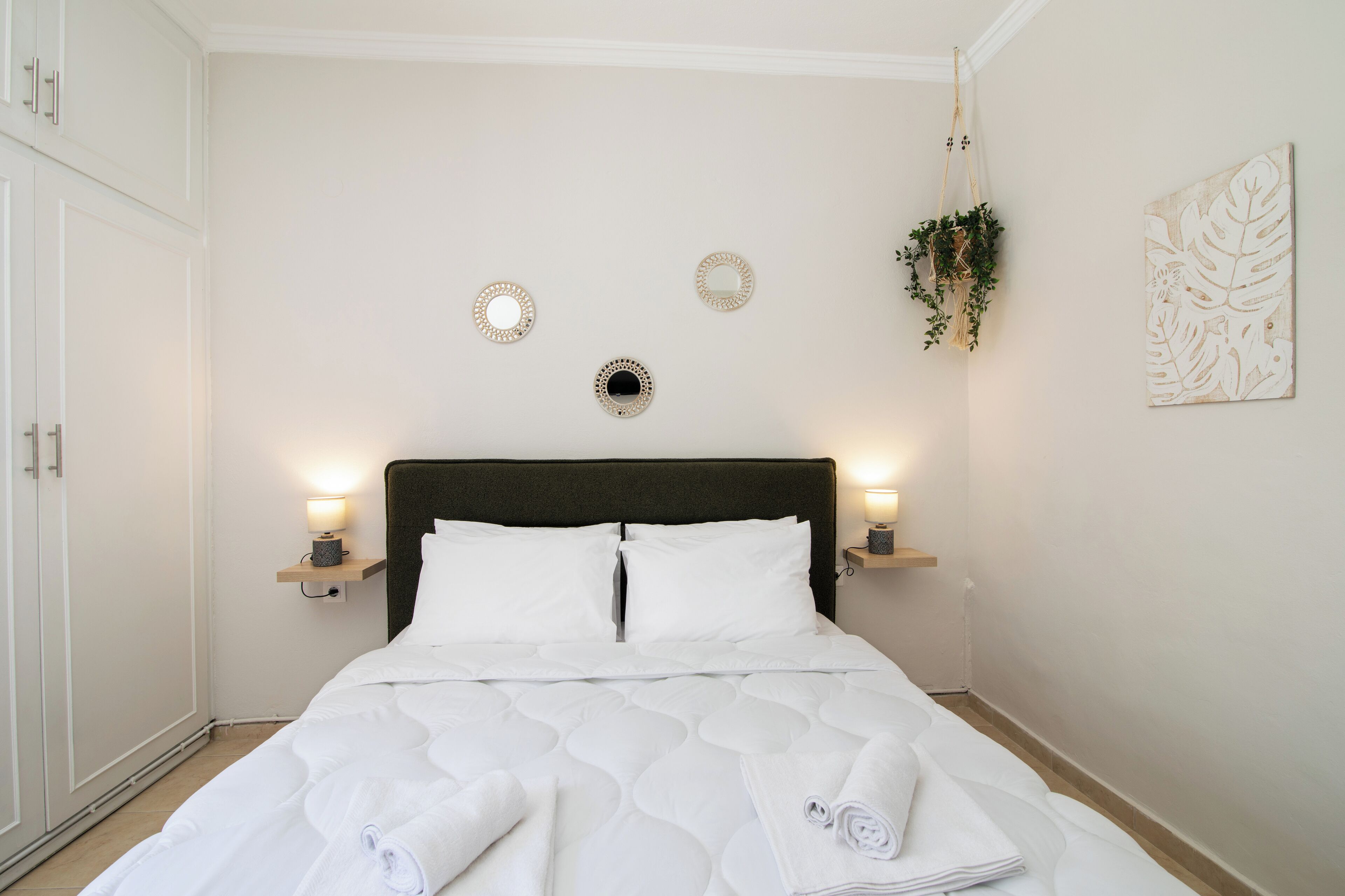 A neatly made bed with white linens and rolled towels, a dark headboard, and wall-mounted sconces with lights.
