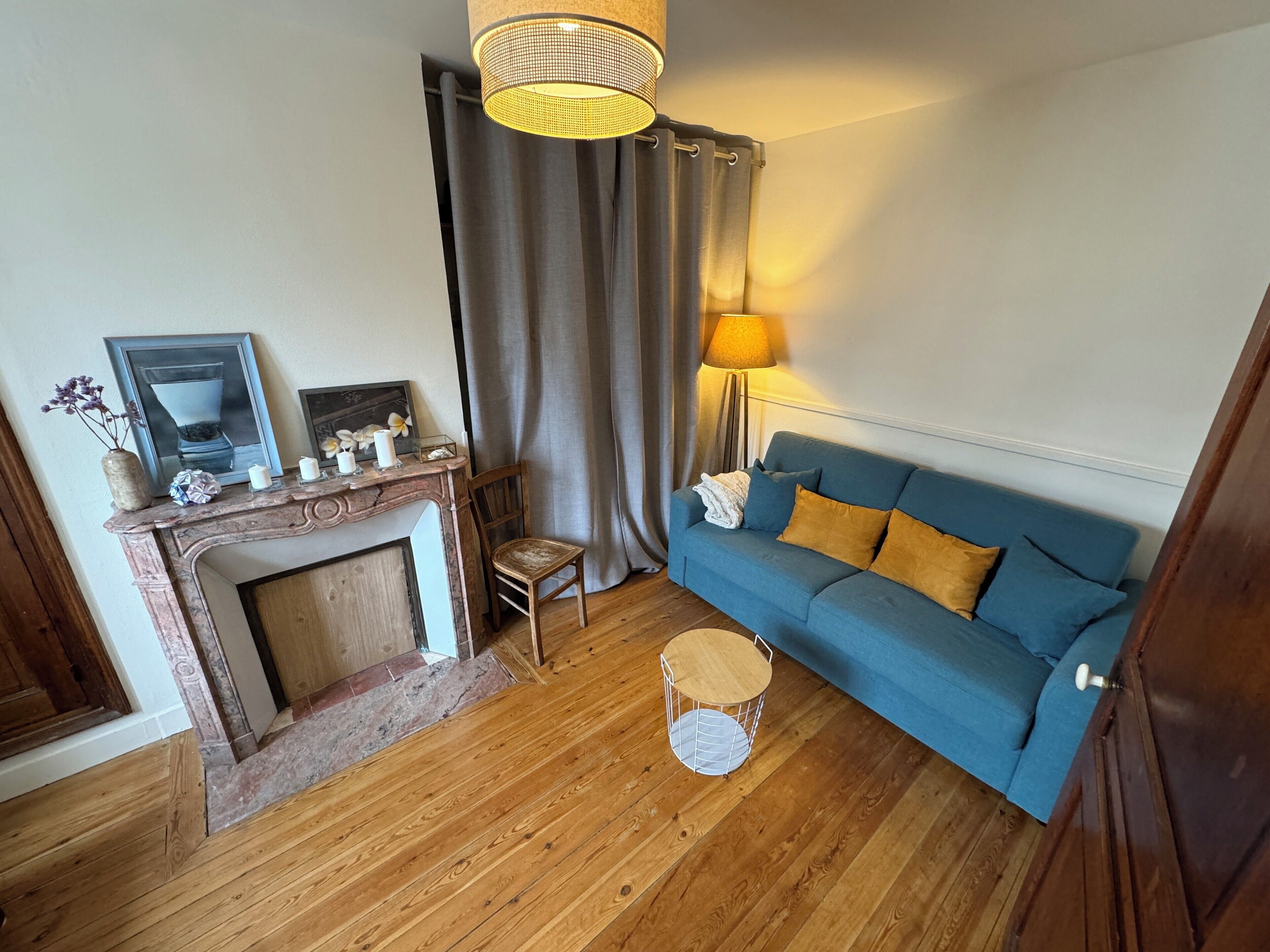 Un salon avec un canapé bleu, une petite chaise en bois, une table ronde en bois, une cheminée et une lampe.