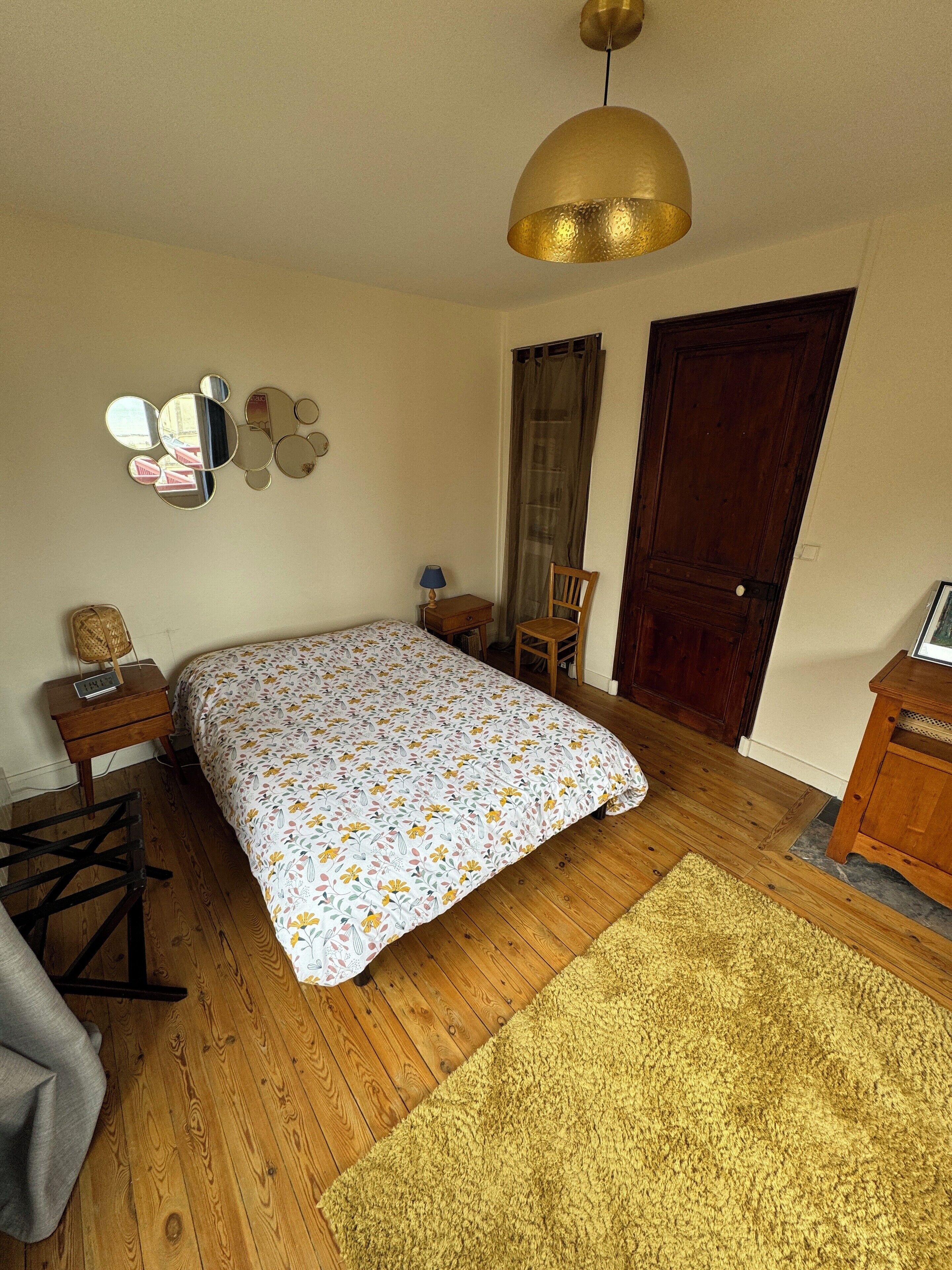 Een slaapkamer met een houten vloer, een bed met een bloemenprint, een houten stoel, een klein tafeltje met een lamp, een spiegel aan de muur en een deur.