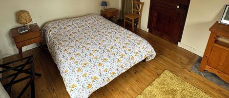 Een slaapkamer met een houten vloer, een bed met een bloemenprint, een houten stoel, een klein tafeltje met een lamp, een spiegel aan de muur en een deur.