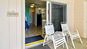 A hotel entrance with a sliding glass door, two white metal chairs, a ceiling fan, and a welcome sign.