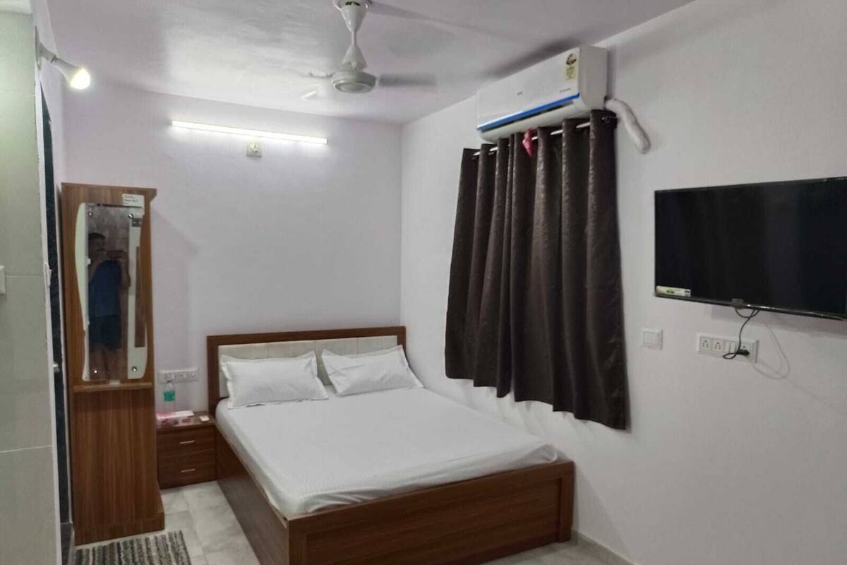 A bedroom with a wooden bed, a ceiling fan, an air conditioning unit, a television, and a mirror.
