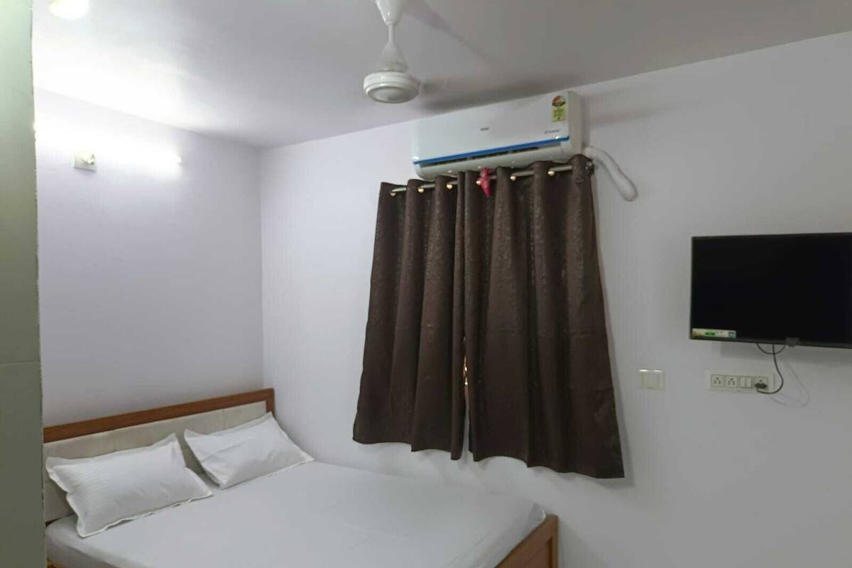 A single bed with white bedding, a ceiling fan, an air conditioning unit, a TV mounted on the wall, and a power outlet.