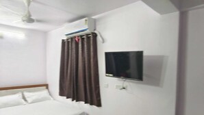 Et soveværelse med seng, ventilator, aircondition, fjernsyn ophængt på væggen og et vindue med gardiner.