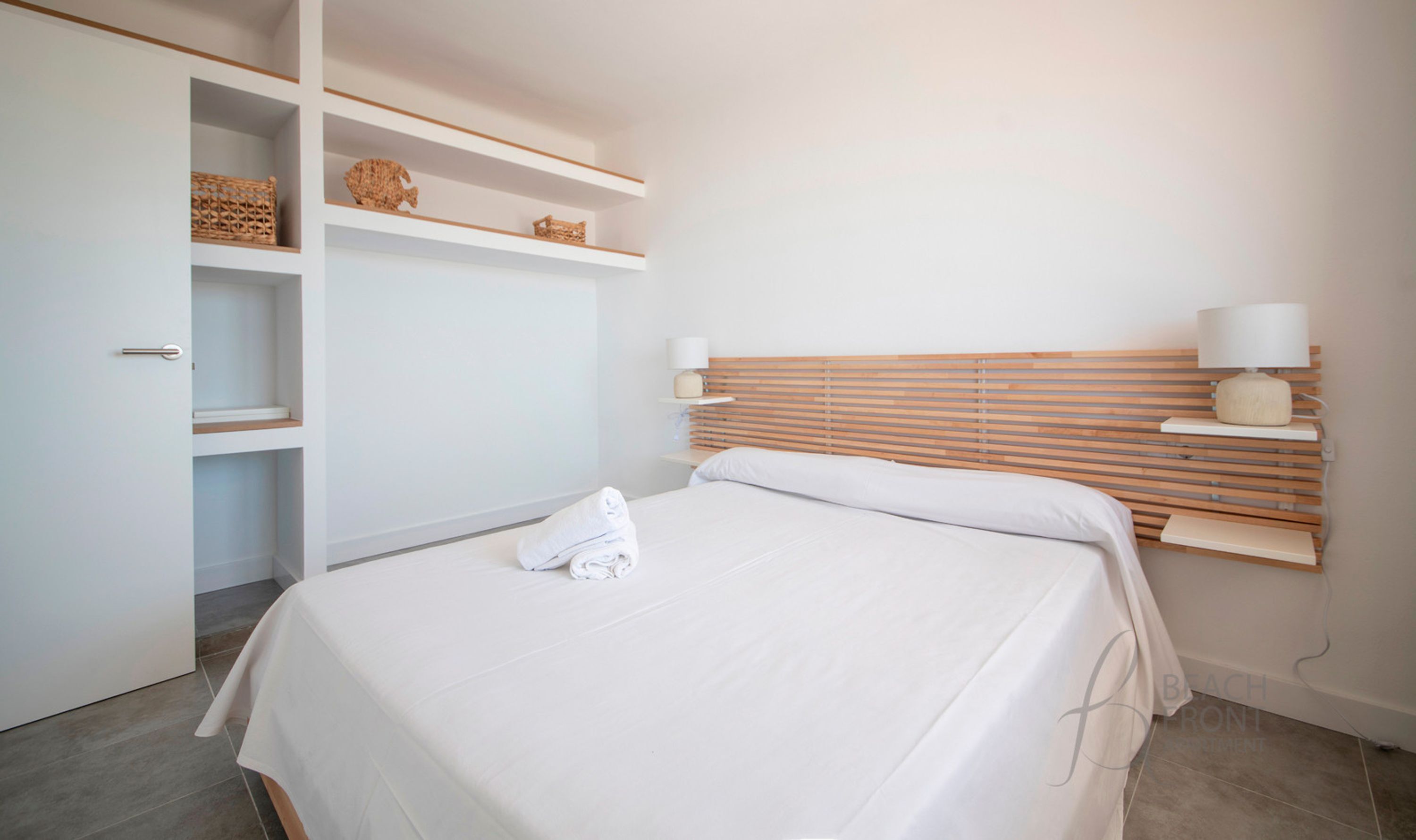 A bedroom with a bed, a wooden headboard, two bedside lamps, and a shelf with decorative items.