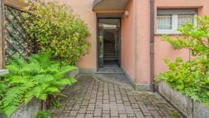 A narrow brick pathway leading to a building entrance with a curved roof, surrounded by lush greenery and a metal fence. - Sunflowers Home - by Varenna Holidays (Mandello del Lario)