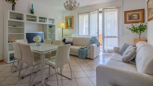 A modern living room with a dining area, white furniture, a chandelier, and a large window with curtains. - Sunflowers Home - by Varenna Holidays (Mandello del Lario)